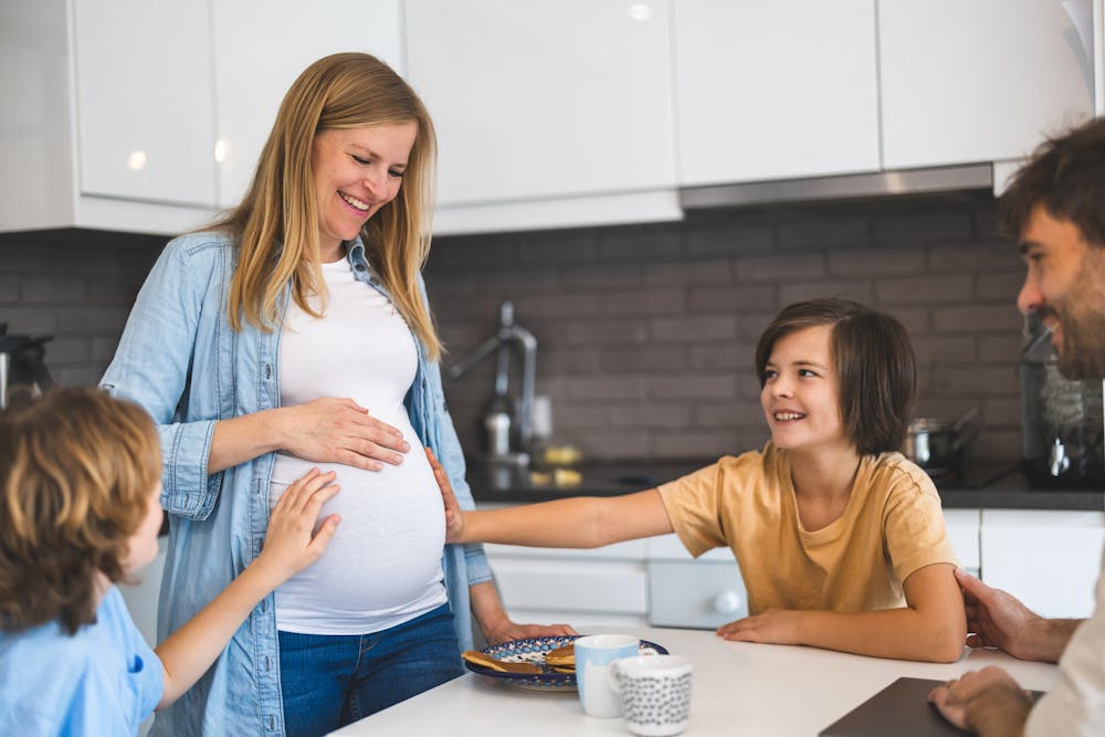 A pregnant woman smiles while a child touches her belly and another looks on; a man observes affectionately, all gathered around a kitchen counter.