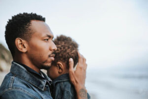 A man gently holds a child, both in denim attire, against a blurred coastal backdrop. The mood appears contemplative and serene.