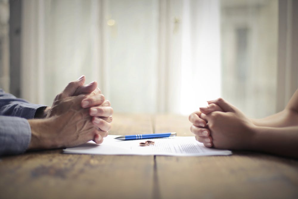 Two pairs of clasped hands rest on a table with a document, pen, and keys, conveying negotiation or agreement, set in a room with soft lighting.