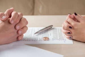 Two people's hands clenched in tension above a document, a pen, and wedding rings, suggesting the context of divorce proceedings on a wooden table.