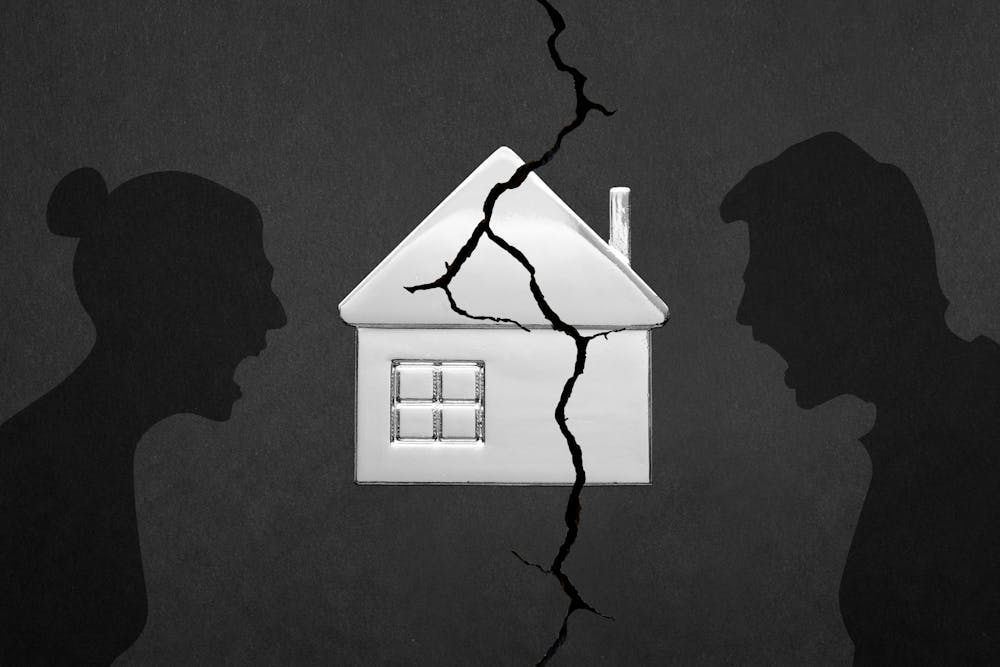 A small white house model with a cracked facade is flanked by silhouettes of a woman's and a man's heads facing away from each other against a gray background.