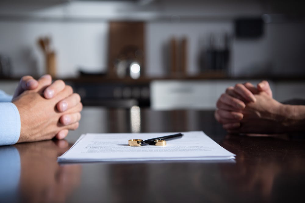 Two people, with hands clasped, face each other across a table with a document and a pen between them, implying a serious discussion or agreement, in a dimly lit room.