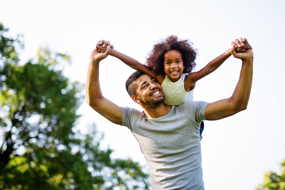 A man lifts a smiling child on his shoulders against a blurry backdrop of greenery. They both appear happy and are outdoors on a sunny day.