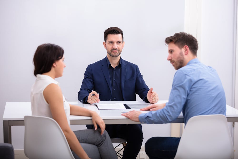 Three people are seated at a table in a meeting setting; one appears to be leading the discussion in a professional office environment. There is no text in the image.