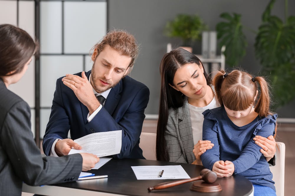 A man in a suit examines documents during a meeting with a woman and child, perhaps in a legal context, indicated by a wooden gavel on the table.