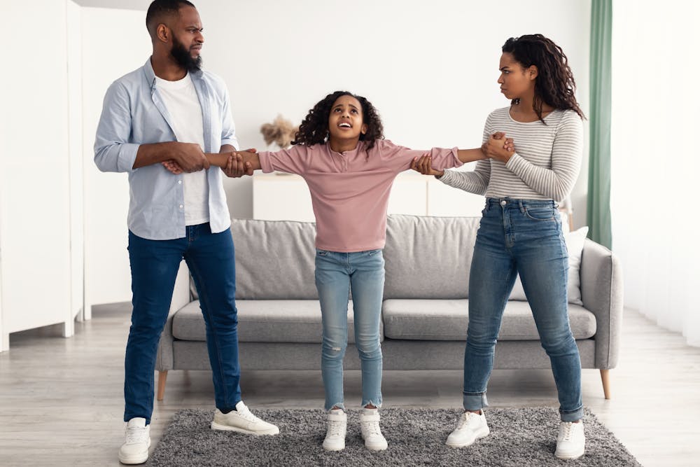 A girl is playfully tugging between a man and a woman in a bright, modern living room, evoking a sense of family togetherness or a lighthearted tug of war.