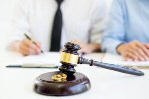 A gavel and two gold wedding rings on a sound block, with two people writing in the background, possibly depicting a legal setting focused on marital issues.