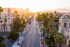 A sunset bathes a cityscape, highlighting palm trees and historical architecture along a wide street, inviting tranquility in a seemingly Californian atmosphere. There is no text in the image.