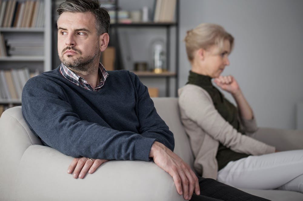 A man and a woman sit back-to-back on a couch, both appearing displeased, in a living room with a bookshelf.