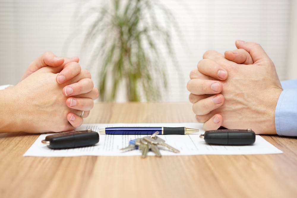 Two people clasp hands on a desk, a pen, keys, and car key fobs between them, suggesting a completed agreement or transaction, possibly for a vehicle.