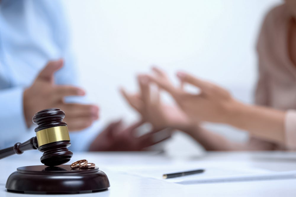 A gavel and two wedding rings lie in focus on a table, with blurred people gesturing in the background, suggesting a divorce or legal proceeding context.