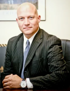 Bald man in a dark pinstripe suit and blue tie sits at a desk with folded hands, wearing a silver watch on his left wrist.